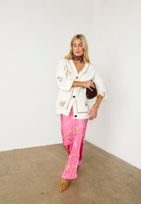 Γυναίκα που φοράει λευκό κεντημένο ζακέτα, ροζ φούστα με σχέδια, καφέ κασκόλ και παπούτσια, κρατώντας ένα σκούρο καφέ φάκελο σε έναν απλό χώρο.