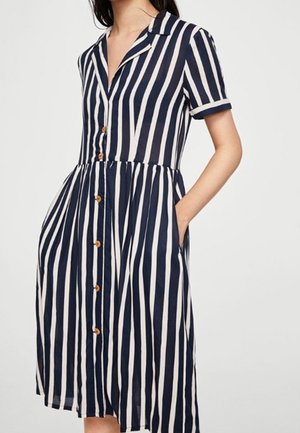 Vrouw draagt een marineblauwe en witte jurk met verticale strepen, korte mouwen, knoopsluiting, kraag en zakken, met een getailleerde taille.