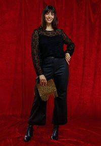 Top en dentelle noire à manches longues avec un motif ondulé, associé à un pantalon noir taille haute et une pochette dorée à perles avec franges.