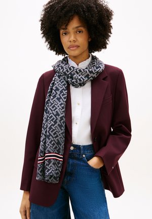 Marineblauwe sjaal met een geometrisch patroon om de nek gewikkeld, gecombineerd met een bordeauxrode blazer en witte blouse, met blauwe spijkerbroek.