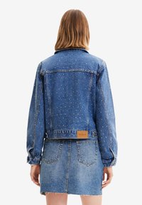 Kvinna som bär en blå denimjacka med flerfärgade små prickar och en matchande denimkjol, visad bakifrån.