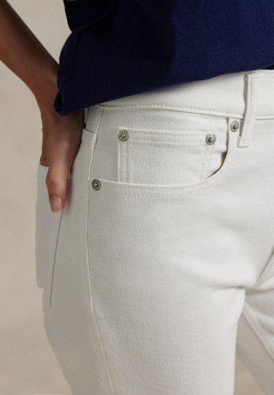 Persoon draagt witte jeans en een marineblauw overhemd, hand rustend op de heup, met zicht op details van het voorzakje en metalen klinknagels.