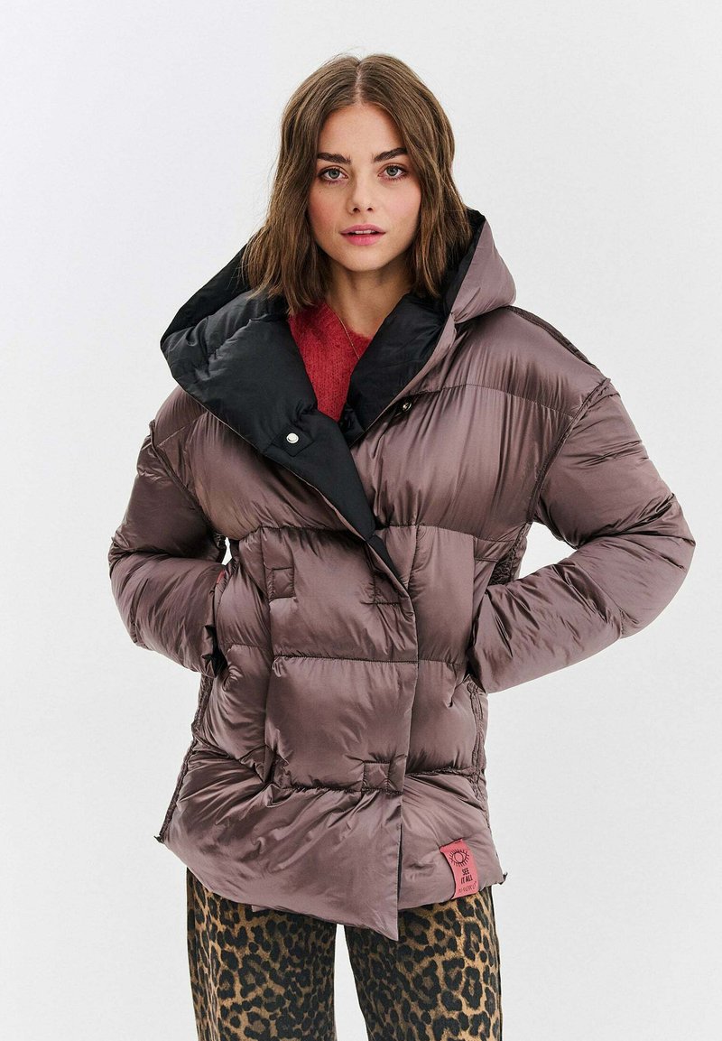 Puchowa, nieregularnie pikowana kurtka w kolorze fioletowym z czarnym kapturem, z zapięciem na guziki i dwiema przednimi kieszeniami. Ozdobiona czerwoną metką.