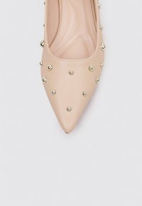 Zapato de tacón nude de cuero con punta afilada, adornado con tachuelas metálicas doradas, que cuenta con una textura suave y un diseño elegante.