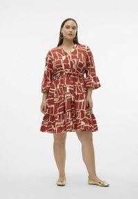 Abito rosso con un motivo geometrico bianco, scollo a V, cintura con fiocco e orlo a balze. Modella che indossa ballerine dorate, mostrando una silhouette ad A.
