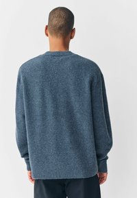 Man die met zijn rug naar de camera staat, gekleed in een loszittende blauwe breisweater met geribbelde manchetten en zoom, in combinatie met donkere broek, tegen een effen achtergrond.