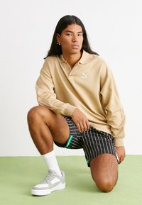 Beige Langarm-Polo mit kleinem weißen Logo, kombiniert mit schwarz gestreiften Shorts und grauen Turnschuhen mit weißen Akzenten und grünen Details.