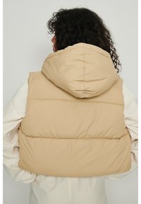 Beżowa pikowana kamizelka z kapturem, o gładkiej fakturze i przeszyciach przy szwach, założona na biały, długi rękaw.