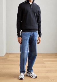 Schwarze Zip-Up-Jacke, blaue Jeans und silberne Sneakers. Der Pullover hat einen gerippten Saum und Bündchen; die Sneakers verfügen über schwarze und silberne Akzente.