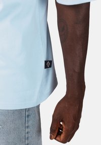 Světle modré tričko s jemnou texturou, které má černou visačku s bílým logem na levé straně poblíž lemu. Detailní záběr ruky, která drží látku.