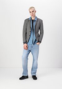 Grauer Blazer über einem Denimhemd, kombiniert mit hellblauen Jeans und schwarzen Sneakers. Das Outfit zeichnet sich durch ein geschichtetes Design mit kontrastierenden Texturen aus.