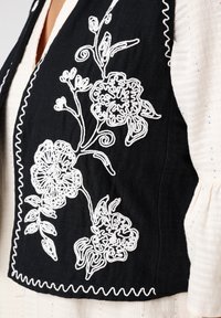 Zwarte geborduurde vest met witte bloemmotieven. Aangevuld met een golvende rand, het is gecombineerd met een licht textiel ondergoed.
