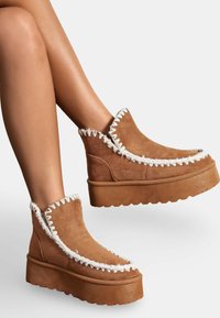 Bottes de cheville en suède marron avec un détail de couture en zigzag blanc le long du bord, dotées d'une semelle plateforme épaisse et d'un design à bout rond.