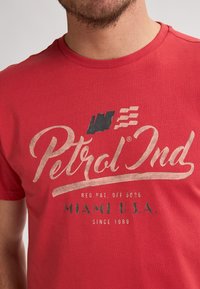 Camiseta de algodón roja con un gráfico grande de estilo vintage que dice "Petrol Ind" en texto crema y verde oscuro, con un logo encima.