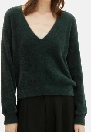 Femme portant un pull à col en V duveteux vert foncé à manches longues et un pantalon noir sur un fond uni.