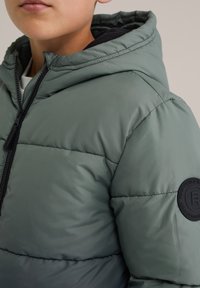 Grön puffjacka med dragkedja fram, svarta detaljer, vadderad design och en rund logotyp-patch på vänster ärm.