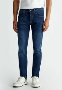 Donkerblauwe spijkerbroek met een slim fit, voorzien van vijf zakken, een knoopsluiting en contrasterende stiksels. Gecombineerd met witte sneakers.