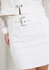 Witte denim mini-jurk met textuur en geometrische patronen, een brede riem met een zilveren gesp en decoratieve drukknoopdetails op de tailleband.