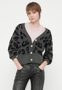Mlada osoba s kratkom kosom nosi sivi kardigan s uzorkom leopard kože s velikim gumbima preko svijetlo ružičaste majice na bretele i crnim teksturiranim hlačama, ruke u džepovima.