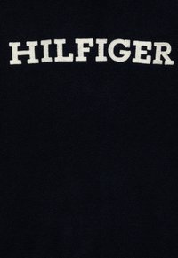 Texto bordado en blanco "HILFIGER" sobre un tejido texturizado azul marino oscuro, centrado y en letras mayúsculas.