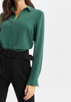 Femme portant un chemisier vert profond à manches longues, rentré dans un pantalon noir taille haute avec une large ceinture et des bagues dorées aux doigts.