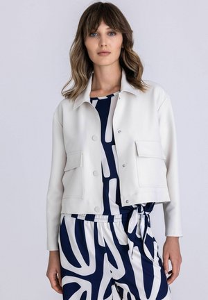 Frau trägt eine weiße kurze Jacke über einem marineblauen und weißen abstrakt gemusterten Kleid, das in der Taille gebunden ist, und steht vor einem schlichten Hintergrund.