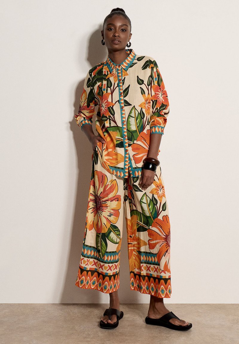 Femme portant une chemise à manches longues et un pantalon large à imprimé floral assortis en orange, vert et beige, avec des sandales noires à enfiler et des bracelets.