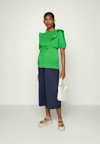 Top verde com pregas e mangas bufantes, com laço na cintura, combinado com culottes azul-marinho. Usado com sandálias de plataforma e uma handbag branca.