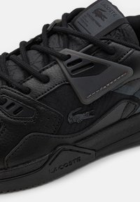 Baskets Lacoste noires avec panneaux en cuir et tissu, lacets noirs, logo Crocodile sur le côté, semelle et languette marquées.