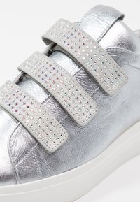 Zilveren leren sneaker met drie grijze klittenbandsluitingen versierd met meerkleurige rhinestones en een witte rubberen zool. Soepele textuur.