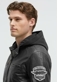 Schwarze Lederjacke mit Kapuze, versehen mit einem Reißverschluss, zwei Fronttaschen und einem Patch am Ärmel. Glatte Textur und taillierte Passform.