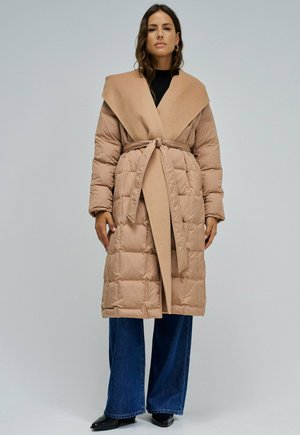 Mujer que lleva un abrigo beige acolchado con cinturón sobre una camiseta negra, vaqueros azules de pierna ancha y zapatos negros, de pie frente a un fondo gris liso.