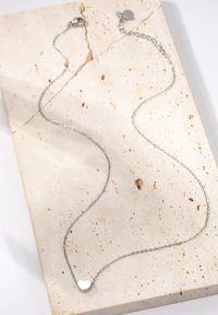 Collier en chaîne argentée avec un petit pendentif en forme de cœur, texture lisse, design minimaliste, présenté sur une surface en pierre de couleur claire.