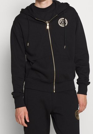 Homme portant un sweat à capuche zippé noir et un pantalon assorti avec des logos Versace Couture dorés sur la poitrine et la cuisse, debout les bras détendus.