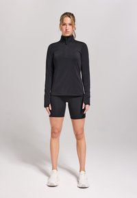 Zwarte lange mouwen sporttop met een kwart ritssluiting, gecombineerd met zwarte fietsshorts en witte sneakers. Glad materiaal, slim-fit ontwerp.