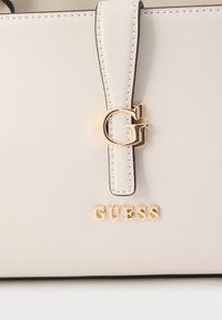 Gaiši bēša rokassomiņa ar gludu ādas apdari, kas izceļas ar zelta toņa akcentiem, izteiksmīgu "G" logotipu un zīmola nosaukumu "GUESS" zelta burtiem.