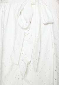 Weißer Baumwollstoff mit bestickten Mustern und Lochdetails. Inklusive eines Stoffbands in der Taille und weichem Textil mit Rüschen.