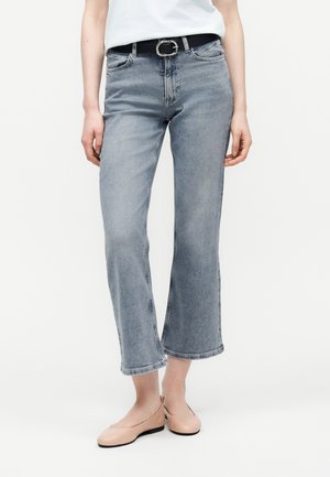 Person trägt ausgewaschene, verkürzte Schlagjeans mit schwarzem Gürtel, weißem Oberteil und beigefarbenen Ballerinas, steht vor einem schlichten Hintergrund.