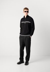 Μαύρο φούτερ με φερμουάρ, υφασμάτινο, με λευκές λεπτομέρειες και λογότυπο. Συνδυασμένο με μαύρο παντελόνι και μαύρα παπούτσια. Τα γυαλιά ηλίου ολοκληρώνουν την εμφάνιση.