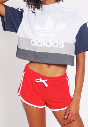 Personne portant un t-shirt court Adidas gris et bleu marine avec logo, et un short de sport rouge avec bordure blanche et cordon, debout avec un bras levé.