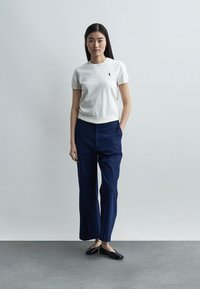 Γυναίκα που στέκεται μπροστά σε απλό γκρι τοίχο, φορώντας λευκό κοντομάνικο πουκάμισο, ναυτικό παντελόνι με φαρδιές πτυχές και μαύρες μπαλαρίνες.