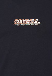 Marineblaues Baumwoll-T-Shirt mit einem auffälligen, stilisierten "GUESS"-Logo in Orange und Blau, das einen leichten Verzerrungseffekt für ein schichtartiges Aussehen aufweist.