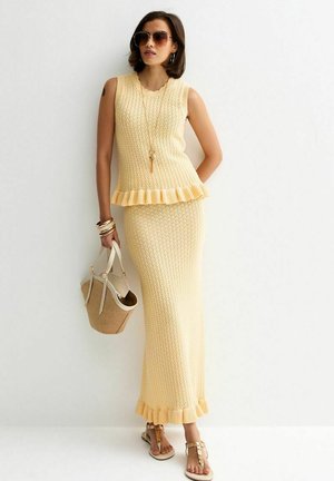 Donna che indossa un top senza maniche giallo con texture e una gonna lunga con bordi arricciati, occhiali da sole, sandali, braccialetti e tiene una borsa beige.