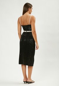Vestido negro con lentejuelas, con un torso ajustado, tirantes finos y una falda a la altura de la rodilla. Presenta un cinturón blanco y un detalle de cremallera en la parte posterior.