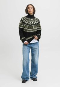 Frau trägt einen schwarzen und olivgrün gemusterten Rollkragenpullover, helle blaue weite Jeans, schwarze Schuhe und steht vor einem einfarbigen Hintergrund.