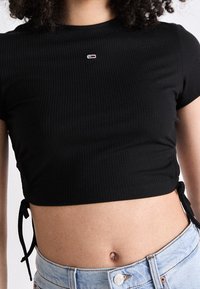 Czarny prążkowany crop top z krótkimi rękawami, z wiązaniami po bokach i małym logo na klatce piersiowej. W połączeniu z jasnoniebieskimi dżinsami z wysokim stanem.