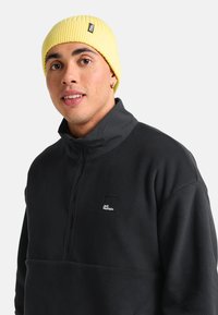 Schwarzer Fleece-Pullover mit hohem Kragen und teilweisem Reißverschluss, versehen mit einem kleinen weißen Logo auf der Brust und kombiniert mit einer gerippten gelben Mütze.
