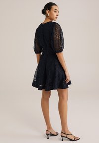 Marinblå spetsklänning med puffärmar, figursydd bodice och utsvängd kjol. Bärs med svarta sandaletter med remmar som visar den delikata spetsmönstret och texturen.