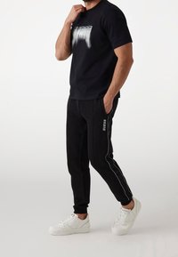 Černé bavlněné tričko s vybledlým bílým potiskem, v kombinaci s černými tepláky s bílými bočními pruhy a bílými teniskami.