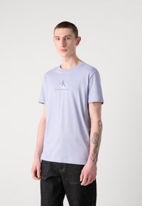 Camiseta de algodón de color morado claro con cuello redondo y mangas cortas, con el logo de Calvin Klein Jeans impreso en la parte delantera. Combinada con pantalones oscuros.